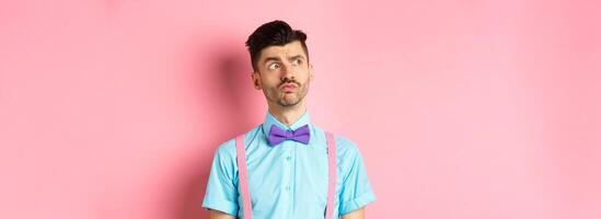 Pensive young man in romantic outfit, looking away and thinking, standing thoughtful on pink background in fancy bow-tie and shirt photo