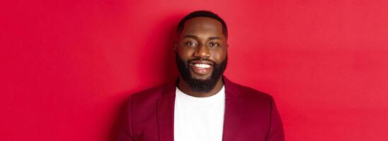 Close-up of handsome and stylish Black man wearing party blazer, smiling happy at camera, standing against red background photo