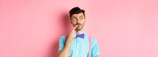 Image of handsome funny man in bow-tie looking thoughtful left and touching lip, thinking what to choose, making decision, standing over pink background photo