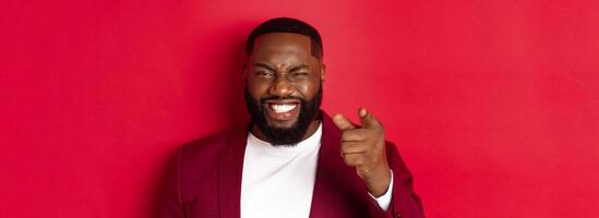 Cheerful Black man with beard wearing party clothes, pointing finger at camera and winking, congratulate with christmas and new year, standing over red background photo