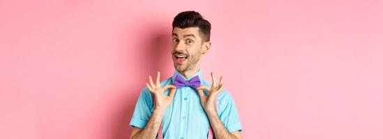 Classy handsome man fixing bow-tie on neck and smiling, getting ready for date or party, standing on pink background photo