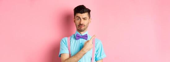 Childish young man acting sad and fake crying while pointing finger upper left corner, complaining on something, standing over pink background photo