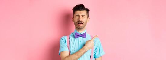 Sad and lonely man in bow-tie crying and pointing finger left, feeling miserable and complaining, sulking while standing on pink background photo