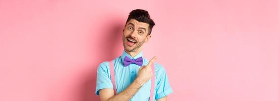Funny male model in bow-tie, tilt head and pointing finger left, inviting to look, check it out gesture, showing advertisement on pink background photo