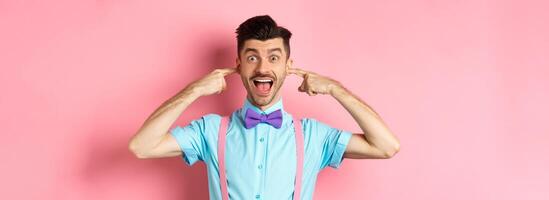 Childish funny guy shut ears and laughing at camera, refuse to listen, standing ignorant on pink background photo