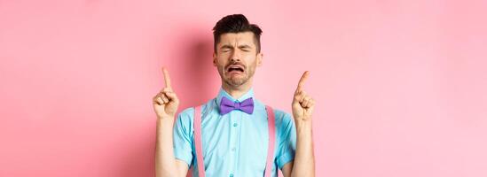 Sad and miserable guy sobbing and crying, pointing fingers up and something disappointed, standing upset on pink background photo