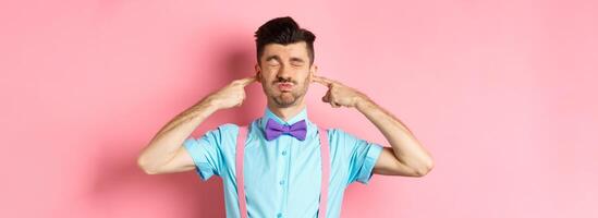 Annoyed funny guy in bow-tie shut ears, plug fingers and grimacing, irritated by loud noise, standing over pink background photo