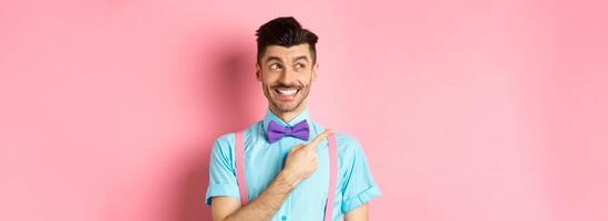 Happy smiling man in funny outfit and bow-tie, pointing finger left and looking at logo, showing advertisement on pink background photo