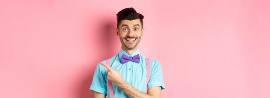Handsome caucasian guy with bristle and moustache pointing finger right aside, showing advertisement, standing over pink background in classy bow-tie photo