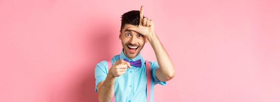 Image of funny guy pointing at you camera and showing loser sign, mocking person who lose, standing over pink background photo