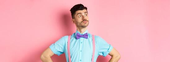 Funny caucasian man with moustache and bow-tie, standing confident with hands spread sideways, looking left at logo proud, standing on pink background photo