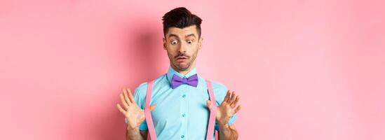 Funny guy with moustache and bow-tie, adjusting his suspenders and looking down with confused and surprised face, standing over pink background photo