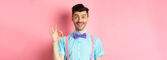 Handsome caucasian man with moustache smiling and showing okay sign, approve something good, praising excellent choice, standing amused on pink background photo