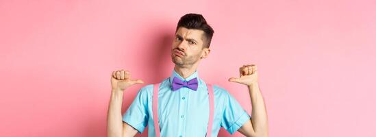 Serious and funny young man trying look cool, pointing at himself to self-promote, being a professional, standing over pink background photo