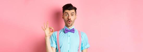 Impressed guy in bow-tie and suspenders saying wow, showing okay gesture with amazement, praise awesome thing, standing over pink background photo