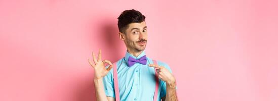 Image of young handsome guy in bow-tie and holiday outfit, pointing at himself and showing okay gesture, self-promoting on pink background photo