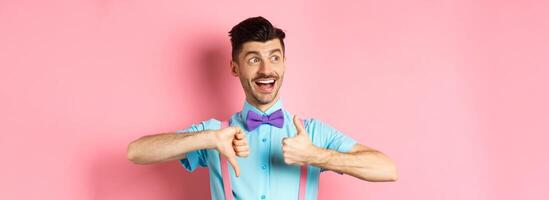 Cheerful man looking happy left, showing thumbs up down, judging product, give positive and negative feedback, standing over pink background photo