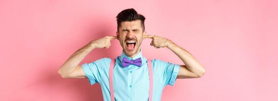 Irritated young man block noise, shut ears with hands and screaming annoyed, being disturbed by loud sound, standing over pink background photo
