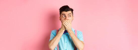 Shocked guy shut mouth, cover lips with hands and looking left speechless, standing in awe over pink background photo
