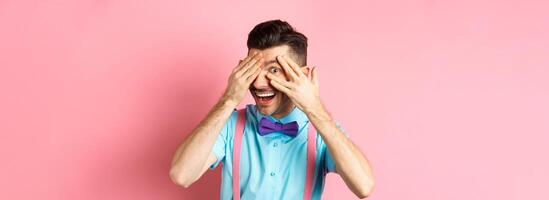 Intrigued funny guy in bow-tie, peeking through fingers at something interesting, checking out something cool, standing on pink background photo