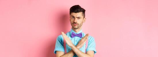 Serious-looking skeptical guy showing cross sign to stop, prohibit something bad, forbid action, standing on pink background photo