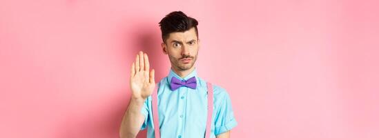 Serious-looking man prohibit something bad, stretch out hand to stop you, forbid action, saying no and looking strict, standing over pink background photo
