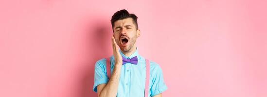 Image of tired guy in bow-tie yawning with opened mouth and closed eyes, standing sleepy, feeling fatigue, standing over pink background photo