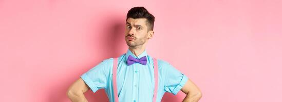 Image of young man with funny moustache and bow-tie standing in confident pose, staring with disbelief and doubts at camera, posing over pink background photo