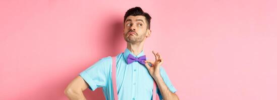 Image of funny man raising chin up and adjusting bow-tie, standing confident and sassy on pink background, looking left photo