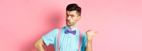 Serious-looking guy in suspenders suggessting to go outside, pointing left and looking confident at camera, starting fight, standing over pink background photo