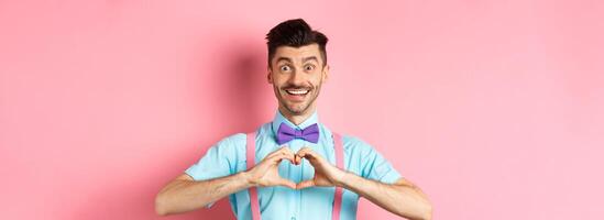 Happy Valentines day. Smiling funny guy with moustache showing heart sign and looking at lover, saying I love you, express feeling, standing over romantic pink background photo
