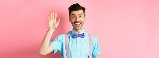 Friendly smiling man in funny bow-tie saying hello, waving hand to greet you, make hi gesture and looking happy yo see you, standing over pink background photo