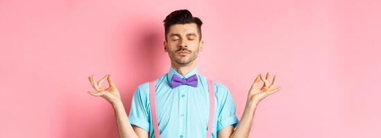 Young guy in bow-tie standing calm and peaceful, meditating with hands in mudra zen gesture and closed eyes, practice yoga to relax, standing over pink background photo