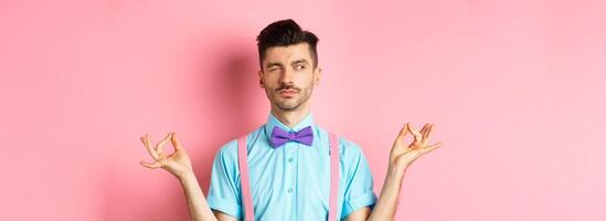 Funny guy with moustache and bow-tie fake meditating, peeking aside while doing yoga asana, standing over pink background photo