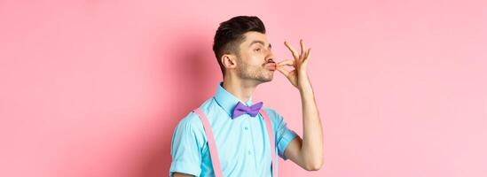 Profile of funny guy touching his french moustache and pucker lips like snob, standing smug on pink background and look left photo