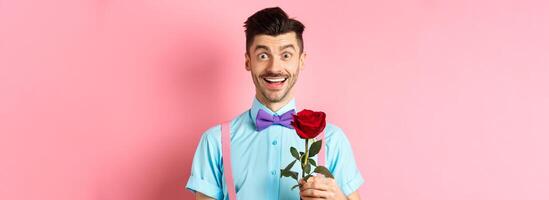 Excited bearded man with moustache and bow-tie waiting for date with red rose, having romantic moment on Valentines day, standing on pink background photo