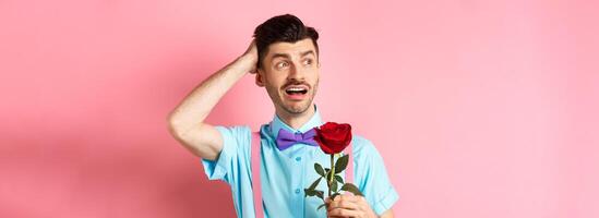 Nervous man waiting for his date on Valentines day, holding red rose and looking confused sideways, scratching head anxiously, standing on pink background photo