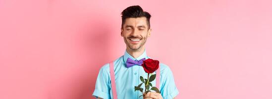 Valentines day and romance concept. Romantic man with red rose going on date with lover, standing in fancy bow-tie on pink background photo