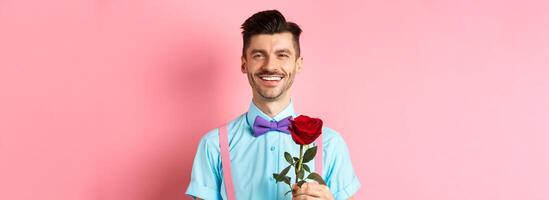 Attractive fancy guy waiting for date on lovers date, holding red rose and smiling, standing on romantic pink background. Valentines day concept photo