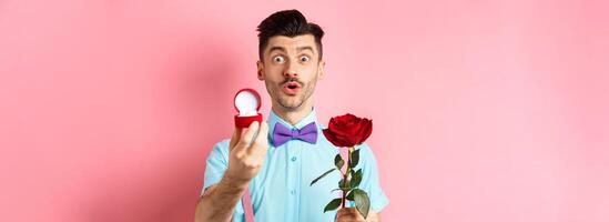 Valentines day. Funny man with moustache and bow-tie making proposal, showing engagement ring and propose with red rose, standing over pink background photo