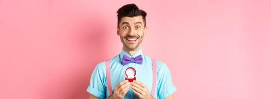 Valentines day. Romantic man in bow-tie showing engagement ring and smiling, asking to marry him, making proposal to lover on pink background photo