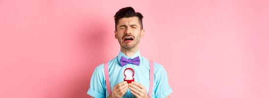 Valentines day. Heartbroken guy crying and holding engagement ring, sobbing from break-up, standing over pink background photo