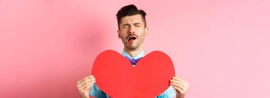 Valentines day concept. Sad and lonely man feeling heartbroken, being rejected, showing big red heart cutout and crying from break-up, standing on pink background photo