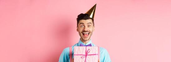 Holidays and celebration concept. Close-up of happy guy in party hat receive birthday gift, scream of joy and cheer, standing over pink background photo