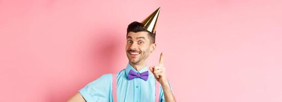 Holidays and celebration concept. Image of happy man in birthday party hat pitching an idea, raising finger and smiling, have plan or solution, standing over pink background photo