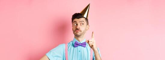 Holidays and celebration concept. Funny guy with moustache and bow-tie, pitch an idea, raising finger and looking aside with silly face, having plan, standing over pink background photo