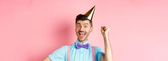 Holidays and celebration concept. Happy birthday boy with moustache and bow-tie celebrating event in party cone hat and festive clothes, raising hand up and shout yes photo