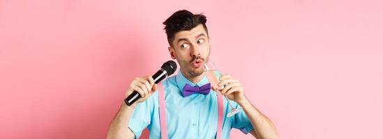 Party and festive events concept. Funny guy singing karaoke, performing song with microphone and drinking champagne from glass, standing on pink background photo