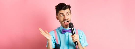 Image of male show host making speech, talking in microphone with clipboard under armpit, enteratin people on festive event, standing over pink background photo