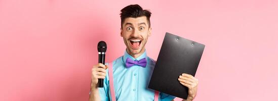 Holidays concept. Happy young man entertainer smiling at camera, holding clipboard and microphone, making speech on party event, standing over pink background photo